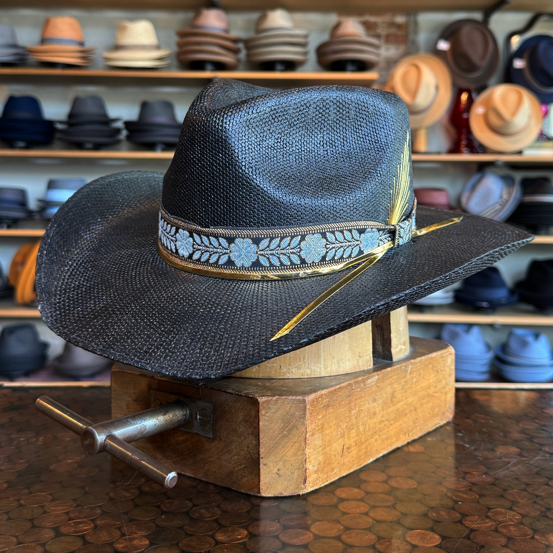 Black woven Wildflower hat with a decorative blue floral band and yellow feather, displayed on a wooden hat block with shelves of various hats in the background.