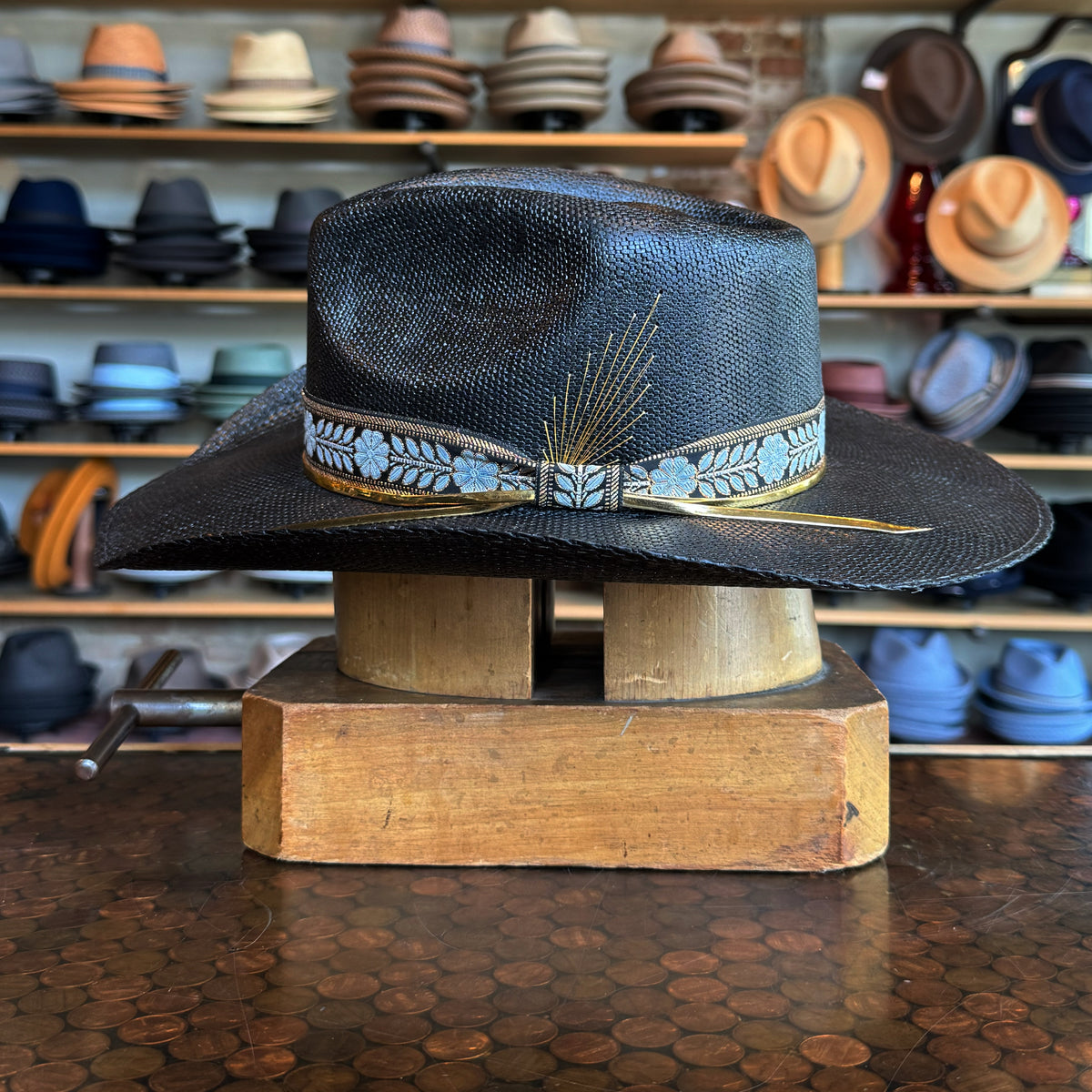 Black Wildflower hat with light blue floral patterned band and decorative feather detail displayed on wooden hat block with various hats in background