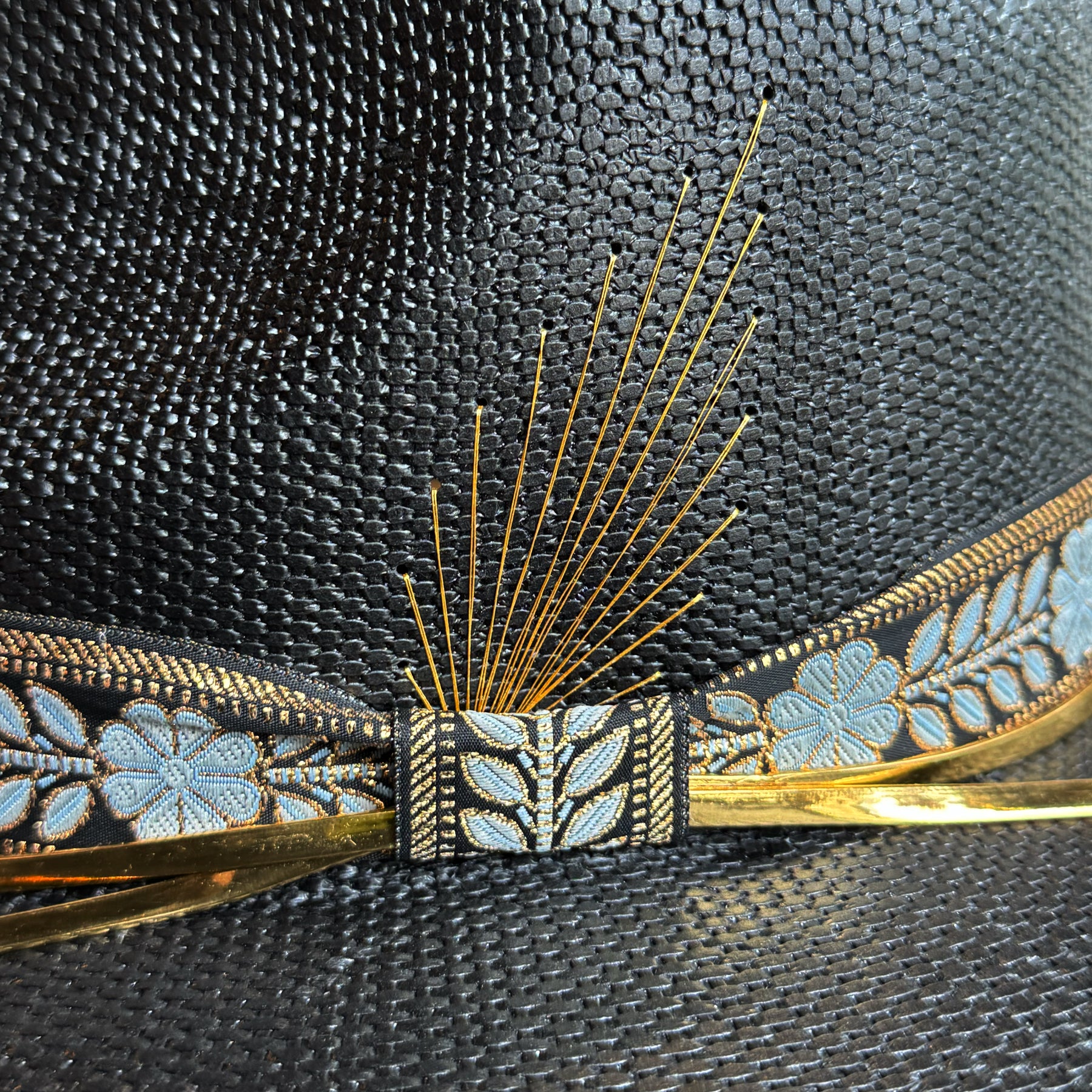Close-up detail of a woven black hat named Wildflower, featuring a decorative band with blue floral embroidery, gold accents, and thin golden spokes arranged like a stylized feather.