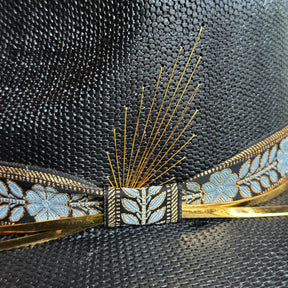 Close-up detail of a woven black hat named Wildflower, featuring a decorative band with blue floral embroidery, gold accents, and thin golden spokes arranged like a stylized feather.