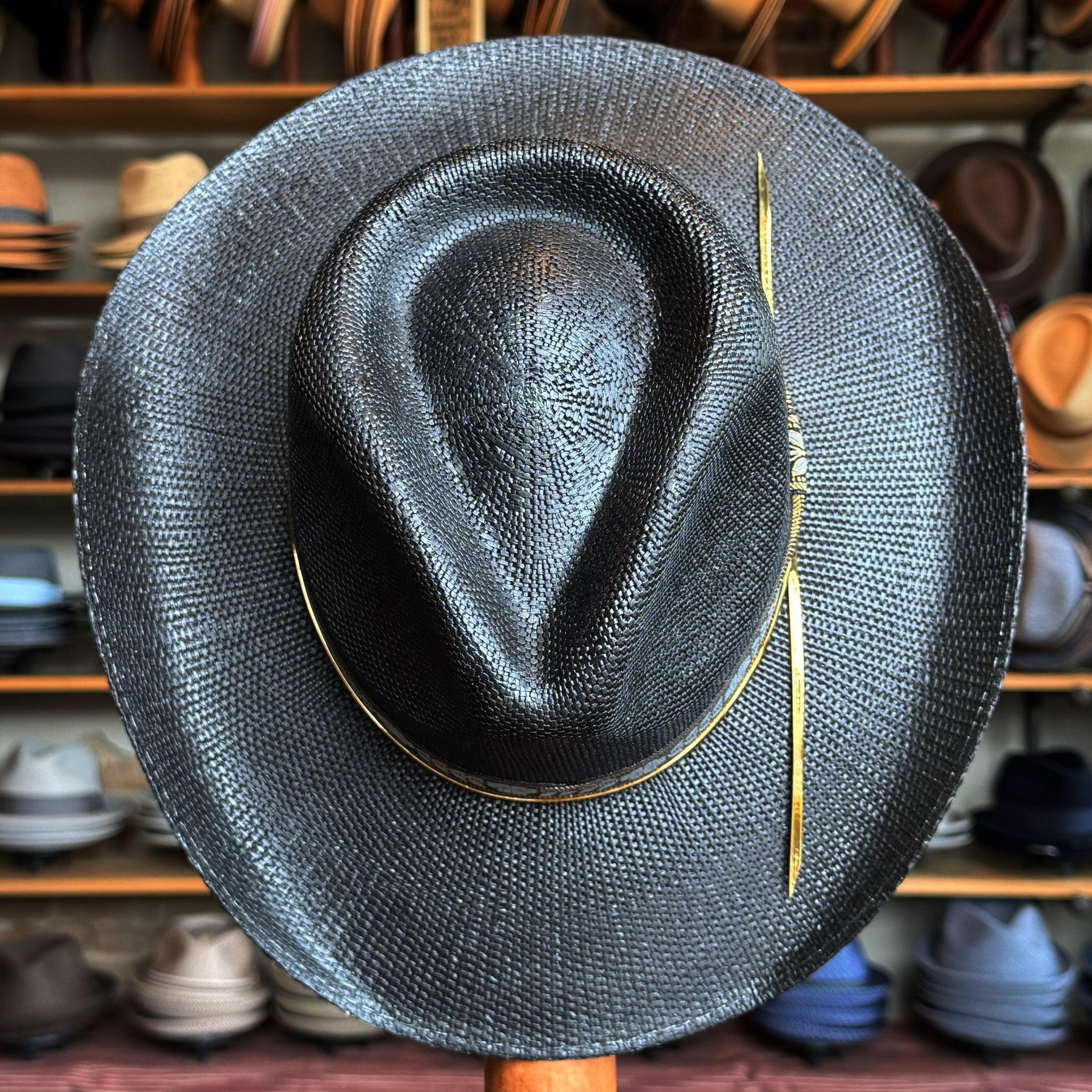 Wildflower black straw brim hat displayed in front of a wall filled with various other hats in a hat shop setting.