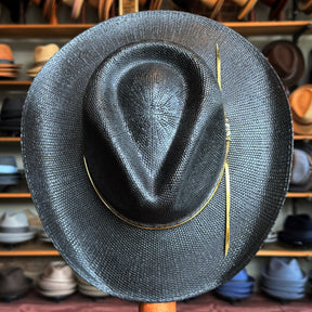 Wildflower black straw brim hat displayed in front of a wall filled with various other hats in a hat shop setting.