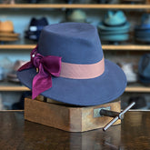 Wool Pinky navy blue fedora hat with a wide taupe band and a burgundy velvet bow accent, displayed on a vintage wooden hat stretcher in a hat shop.
