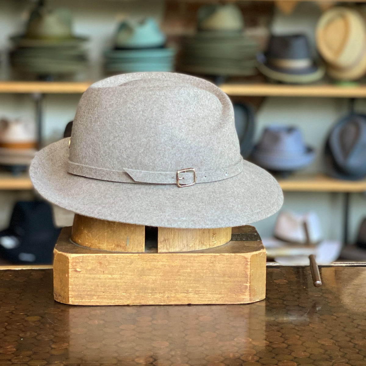 Wool Simone camel-colored wool hat with a soft brim and a thin leather strap and buckle detail, displayed on a wooden stand in a hat shop setting.
