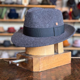 Wool Torino Fedora in charcoal gray with a black band, displayed on a wooden hat stretcher in a store showcasing various hats in the background.
