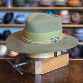 Wormwood Flat Brim Fedora in olive green with a wide flat brim, accented by a matching band and decorative metal clasp, displayed on a wooden hat block in a hat shop.