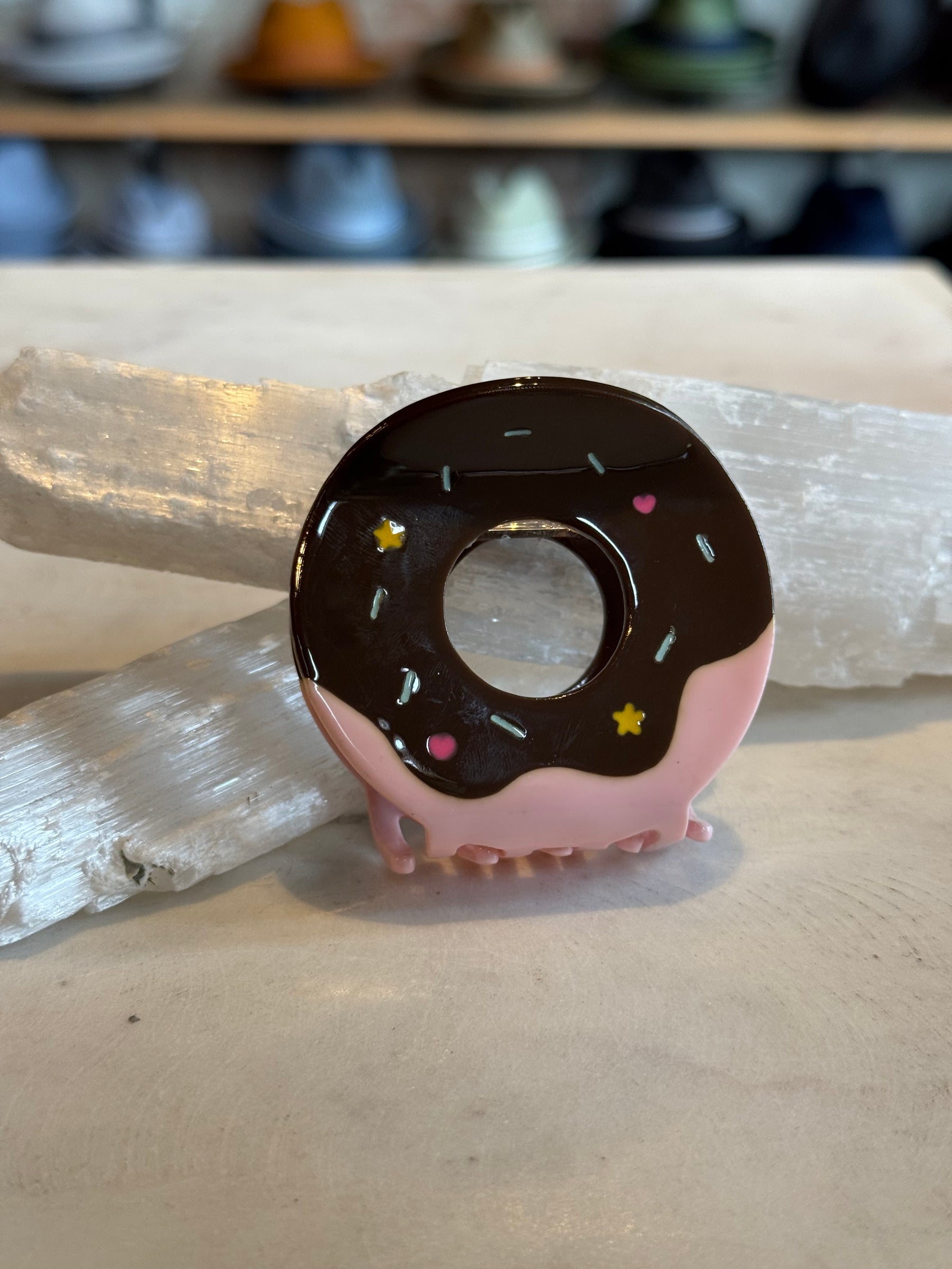 Donut Hair Clip designed with pink and chocolate glaze details, decorated with colorful sprinkles, resting on a neutral surface with a blurred background.