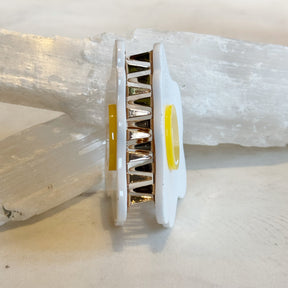 Close-up side view of an Egg Hair Clip showing its white and yellow plastic design with metal clasp details resting against white crystal stones.