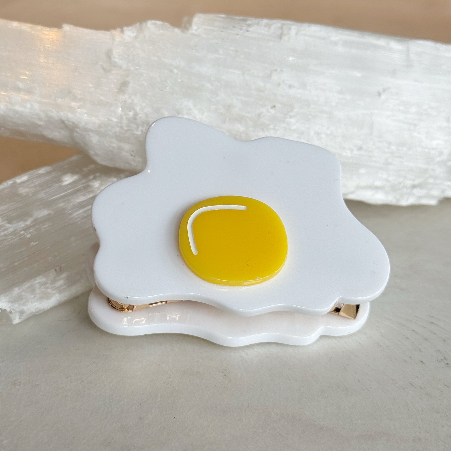 Egg Hair Clip designed to resemble a sunny-side-up fried egg with a bright yellow yolk and white surrounding shape on a neutral background.