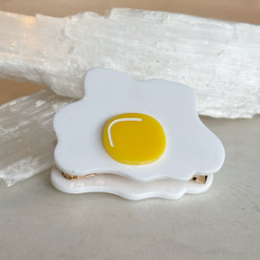 Egg Hair Clip designed to resemble a sunny-side-up fried egg with a bright yellow yolk and white surrounding shape on a neutral background.
