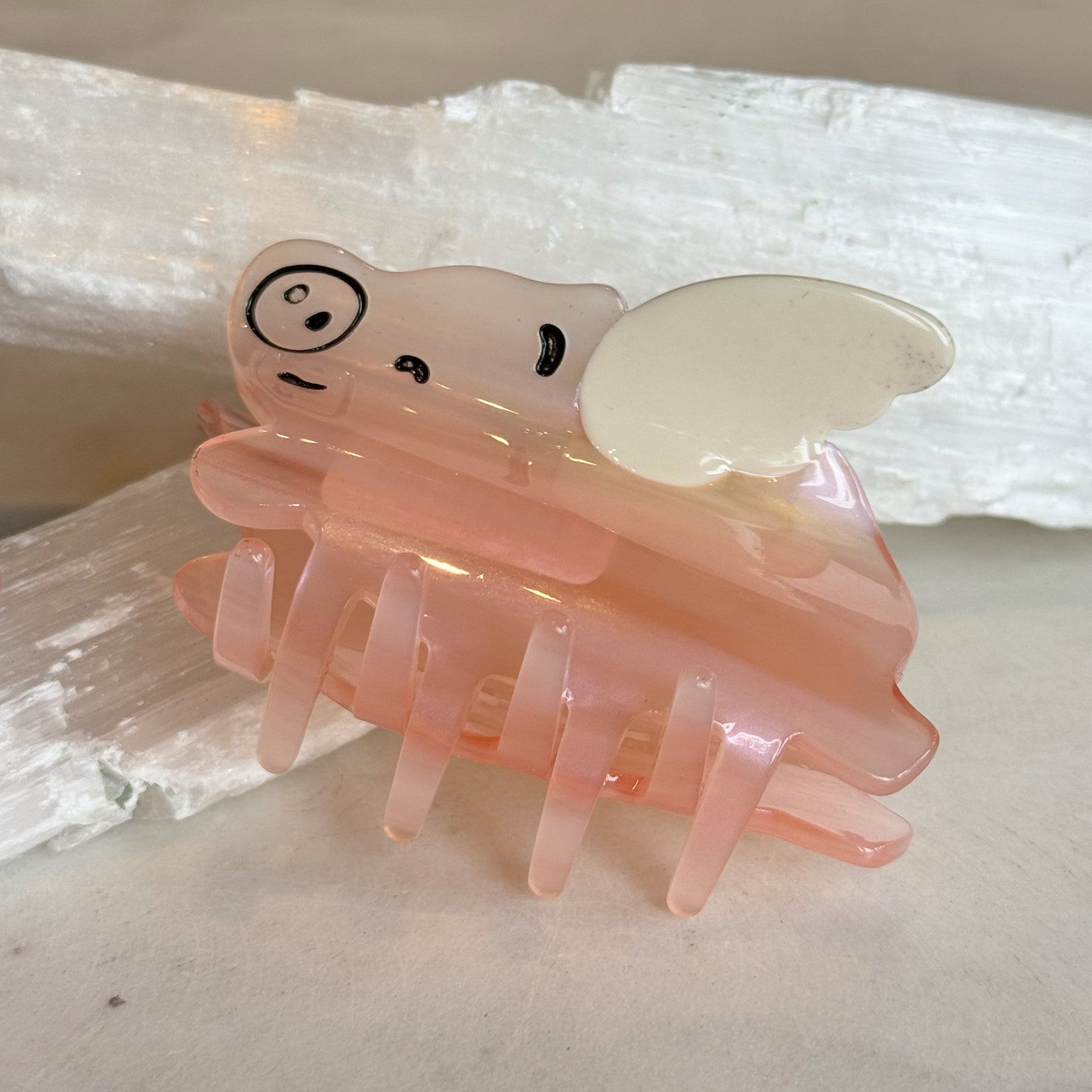 Close-up of a translucent pink Flying Pig Hair Clip with white wings, featuring a playful pig face design, resting on a textured white surface.