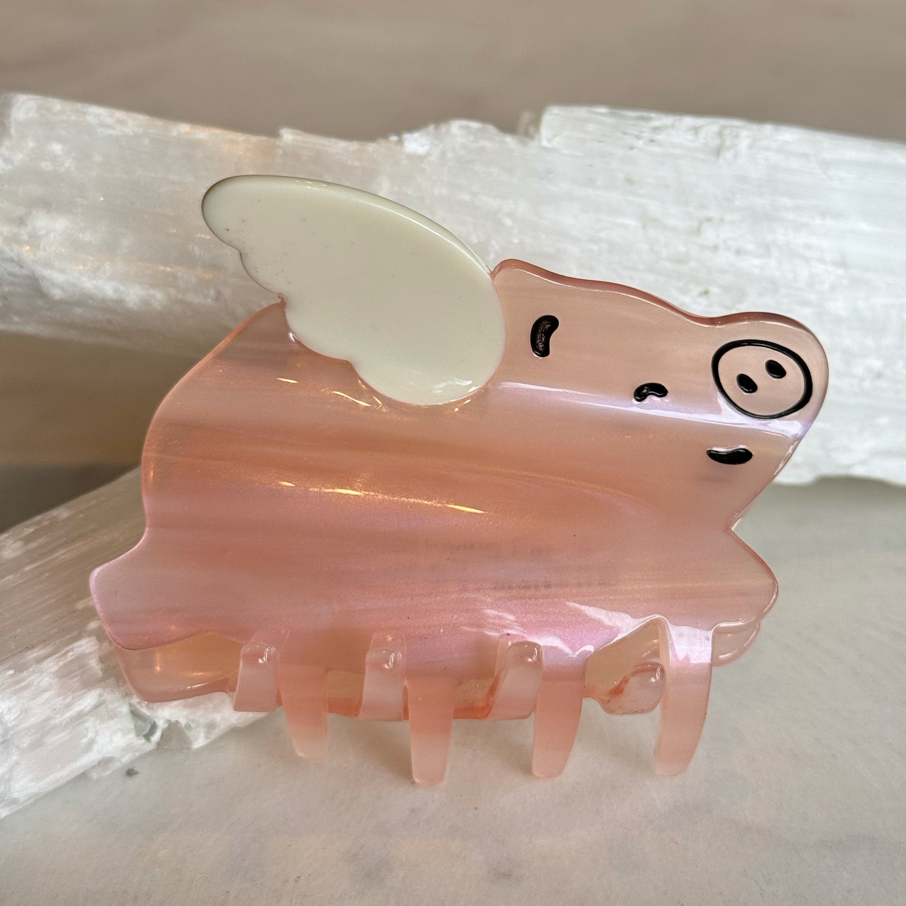 Flying Pig Hair Clip made of glossy pink resin shaped like a pig with white wings and simple black facial features, resting on a light textured surface.