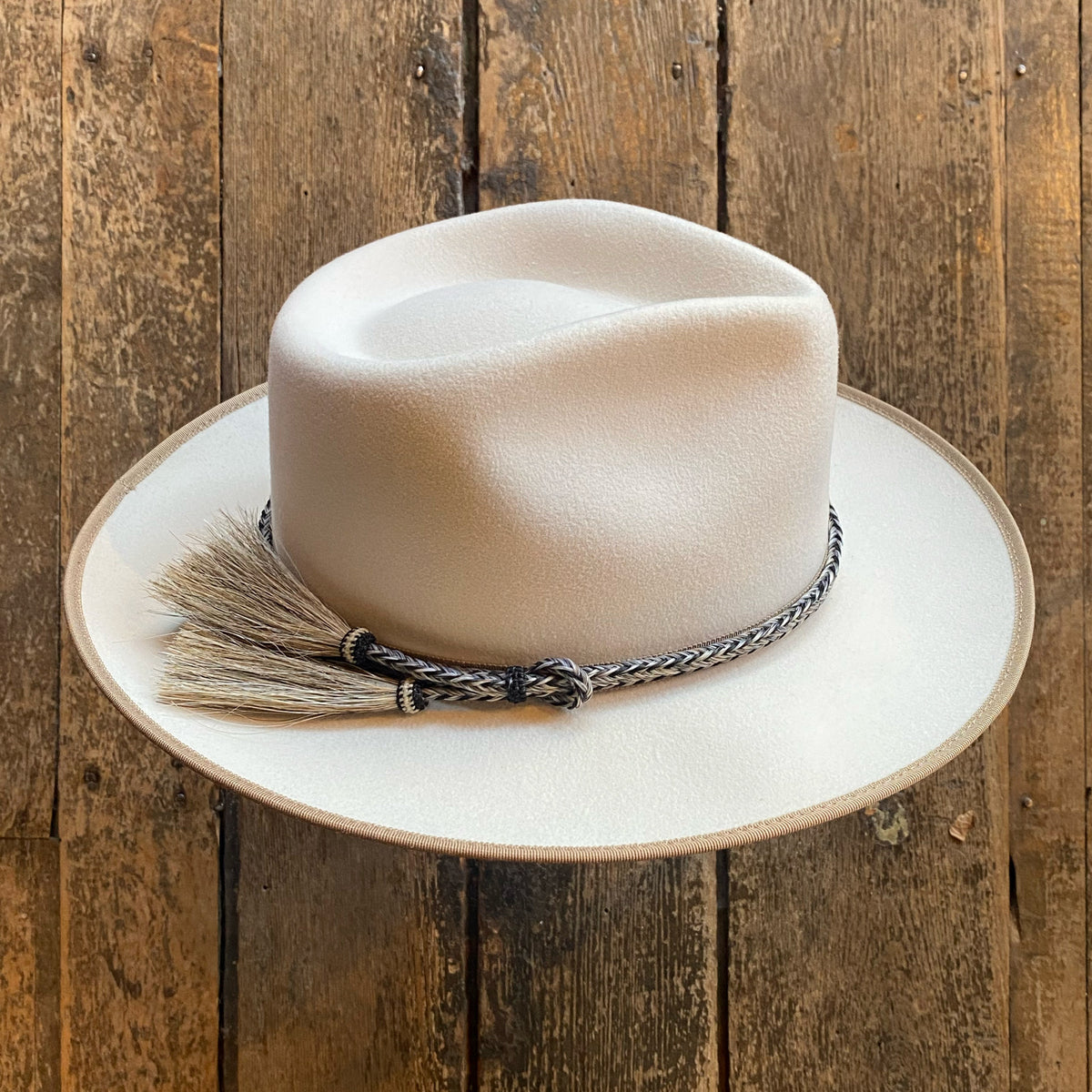Cream-colored fedora hat featuring a 2 Strand Horsehair Hat Band with Side Tassels, displayed on a rustic wooden surface.