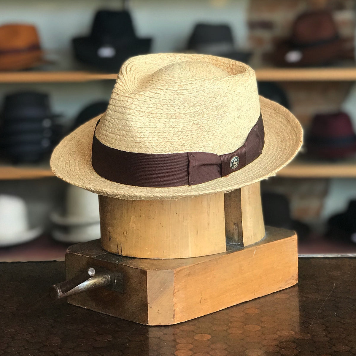 42nd Street natural straw hat with a brown ribbon band displayed on a wooden block against a background of various hats on shelves