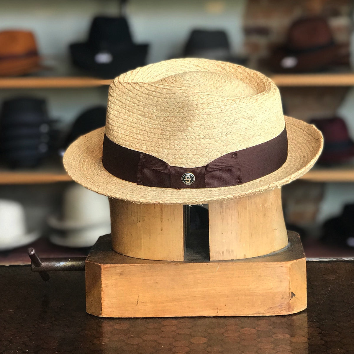 42nd Street straw fedora hat with a dark brown ribbon band and a small branded button, displayed on a wooden hat block in a hat shop.