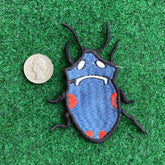 A close-up of an embroidered Insect Patch resembling a blue beetle with red spots and a sad face, placed next to a quarter for size on a green fabric background.