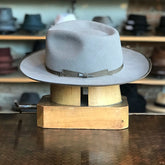 Colver flat brim hat in soft greige color with a matching band and bow detail, displayed on a wooden hat block in a hat shop.