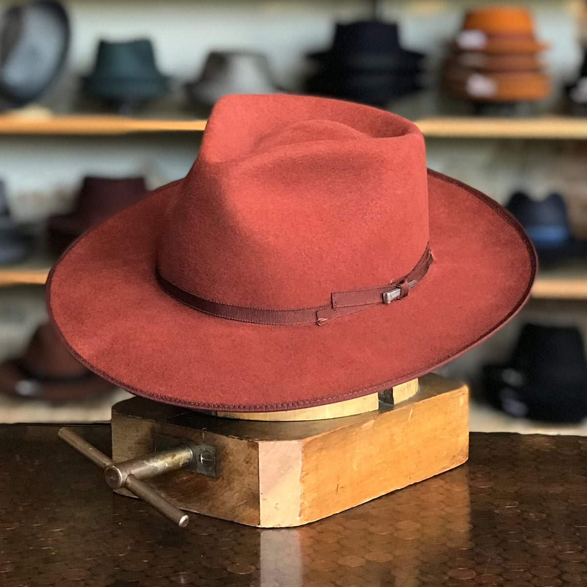 Colver rust-colored felt hat with wide brim and brown leather band, displayed on a wooden stand in a hat shop setting.