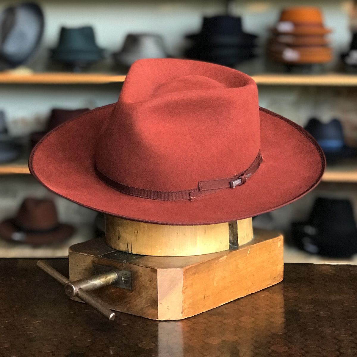 Colver flat brim hat in warm ochre color with a narrow matching band, displayed on a wooden hat block in a shop setting with various hats in the background.