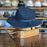Elam black felt fedora hat with a sleek leather band detail, displayed on a wooden hat block in a vintage-style hat shop setting.
