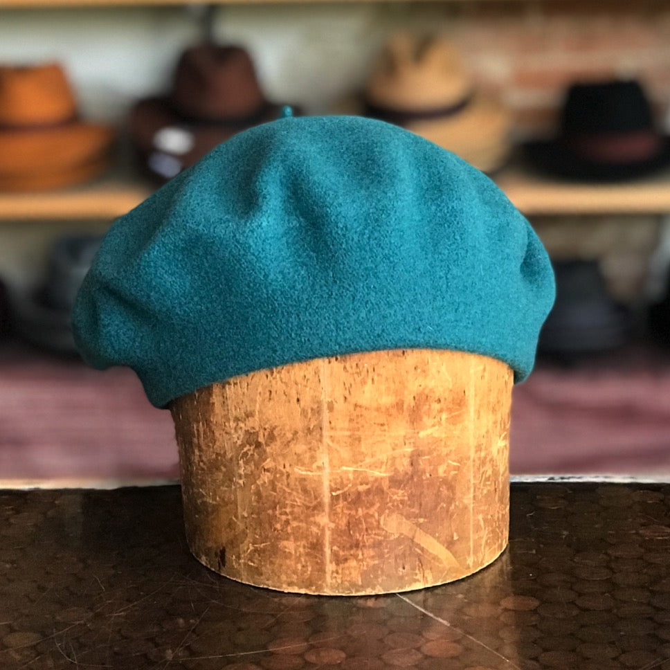 Front of Teal wool Flora Beret displayed on a wooden stand with various hats blurred in the background, showcasing a classic and stylish accessory.