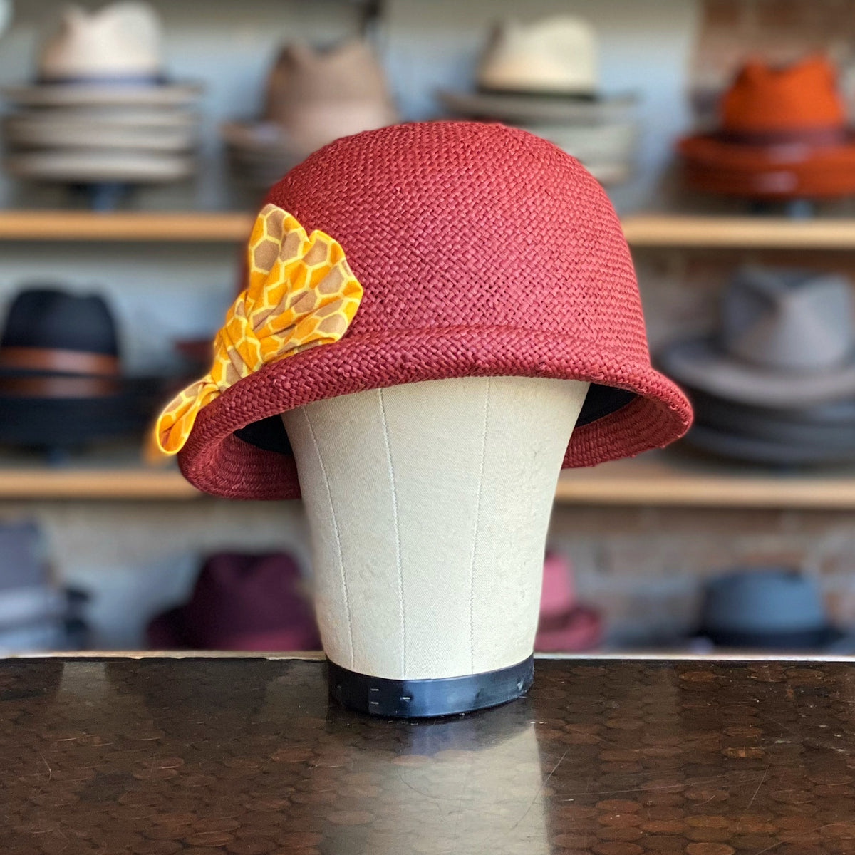 Happy Talk Cloche in brick red with a textured finish and a decorative yellow honeycomb-patterned fabric accent on the side, displayed on a mannequin head.