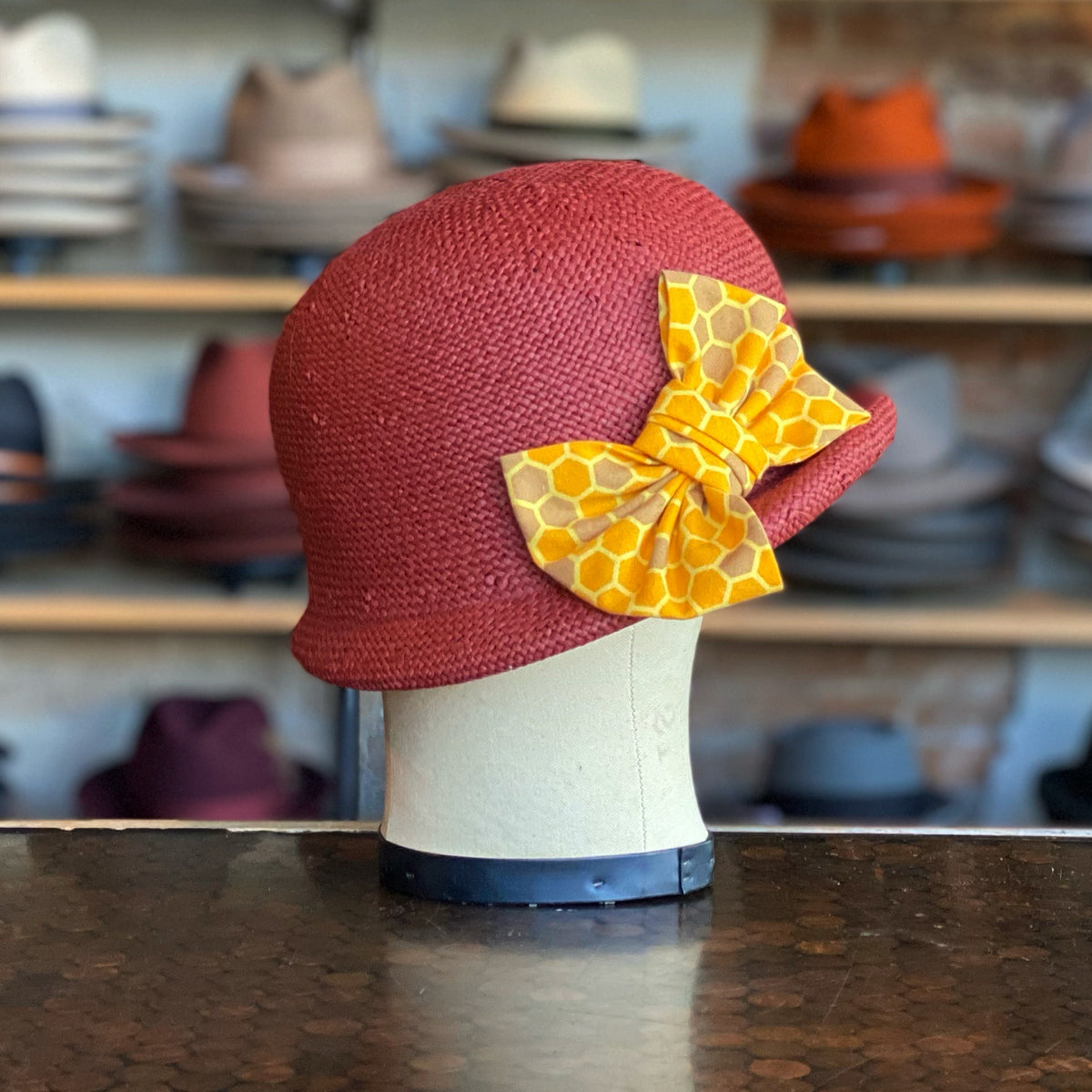 Happy Talk Cloche in a textured brick red fabric featuring a yellow honeycomb patterned bow, displayed on a mannequin head with a background of assorted hats.