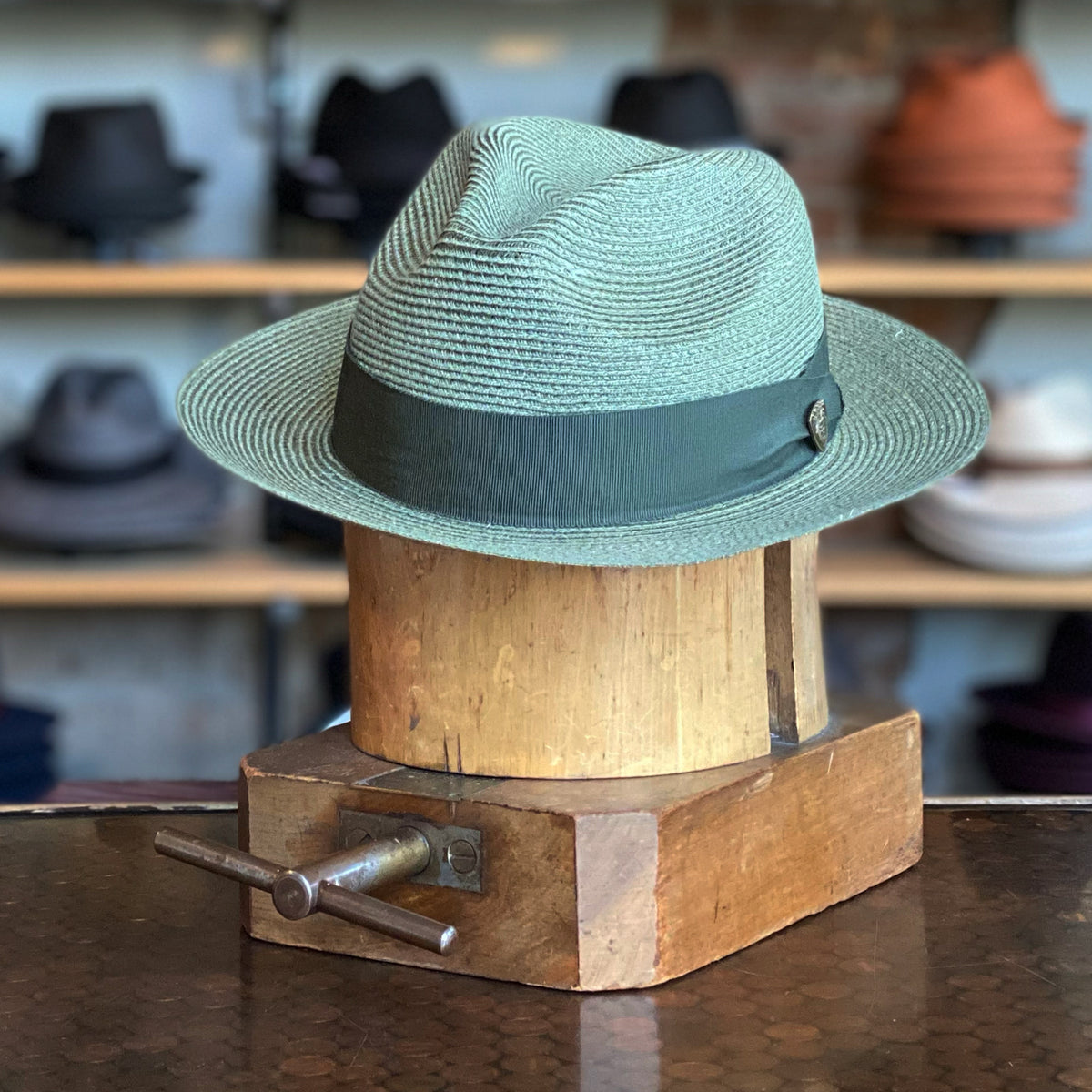 Hemp Rosebud sage green woven flat cap with a dark green fabric band, displayed on a wooden hat block in a hat shop with blurred hats in the background.