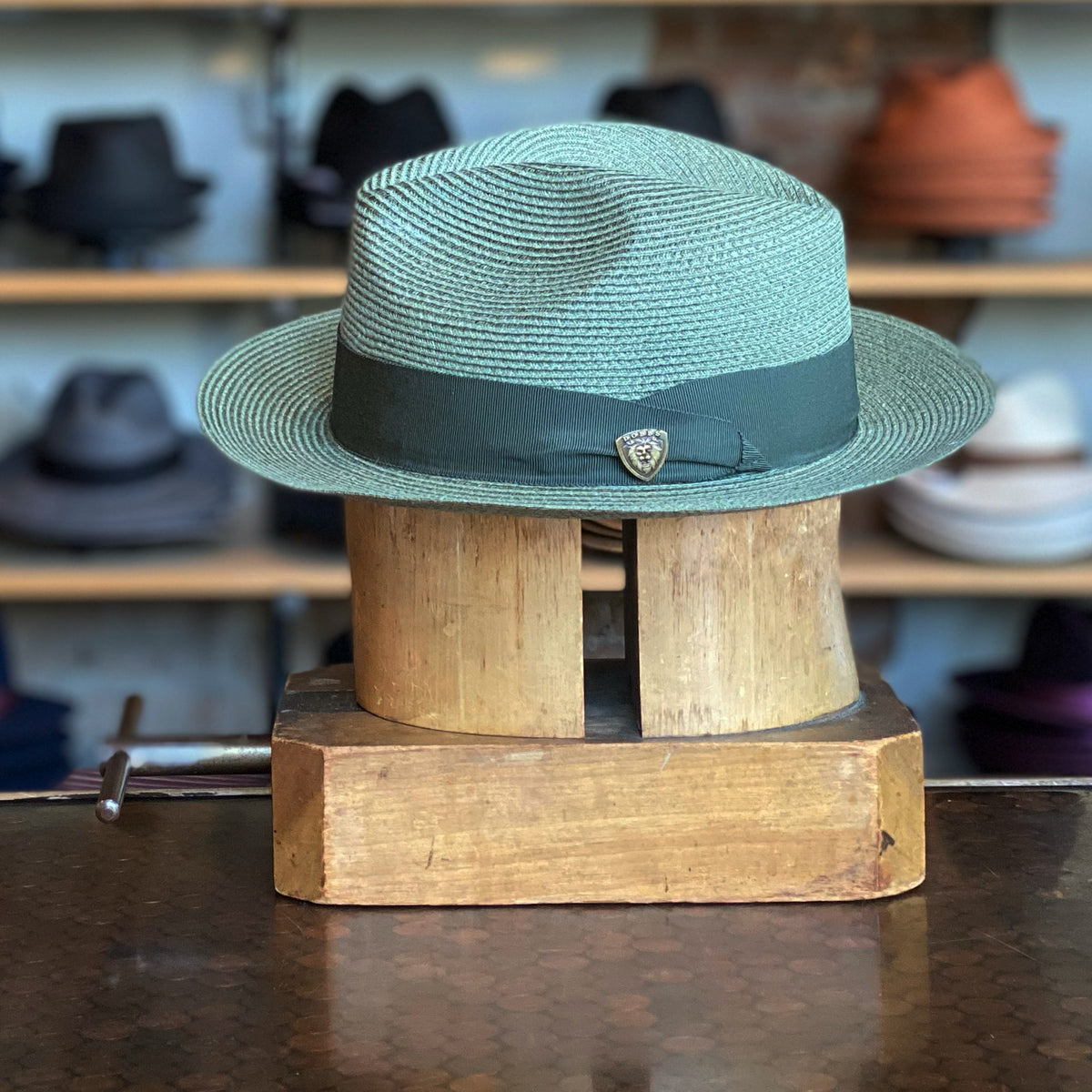 Hemp Rosebud sage-colored woven hat with a dark green band and small decorative pin, displayed on a wooden hat block with shelves of various hats in the background.