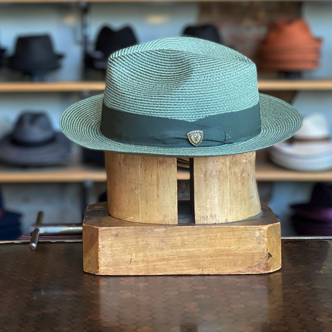 Hemp Rosebud sage-colored woven hat with a dark green band and small decorative pin, displayed on a wooden hat block with shelves of various hats in the background.
