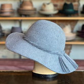 A soft grey felt wide-brimmed hat with decorative tassels on the side displayed on a mannequin head, styled as the Kitten.