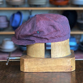 Linen Hatteras burgundy linen flat cap displayed on a wooden hat block with shelves of various hats blurred in the background.