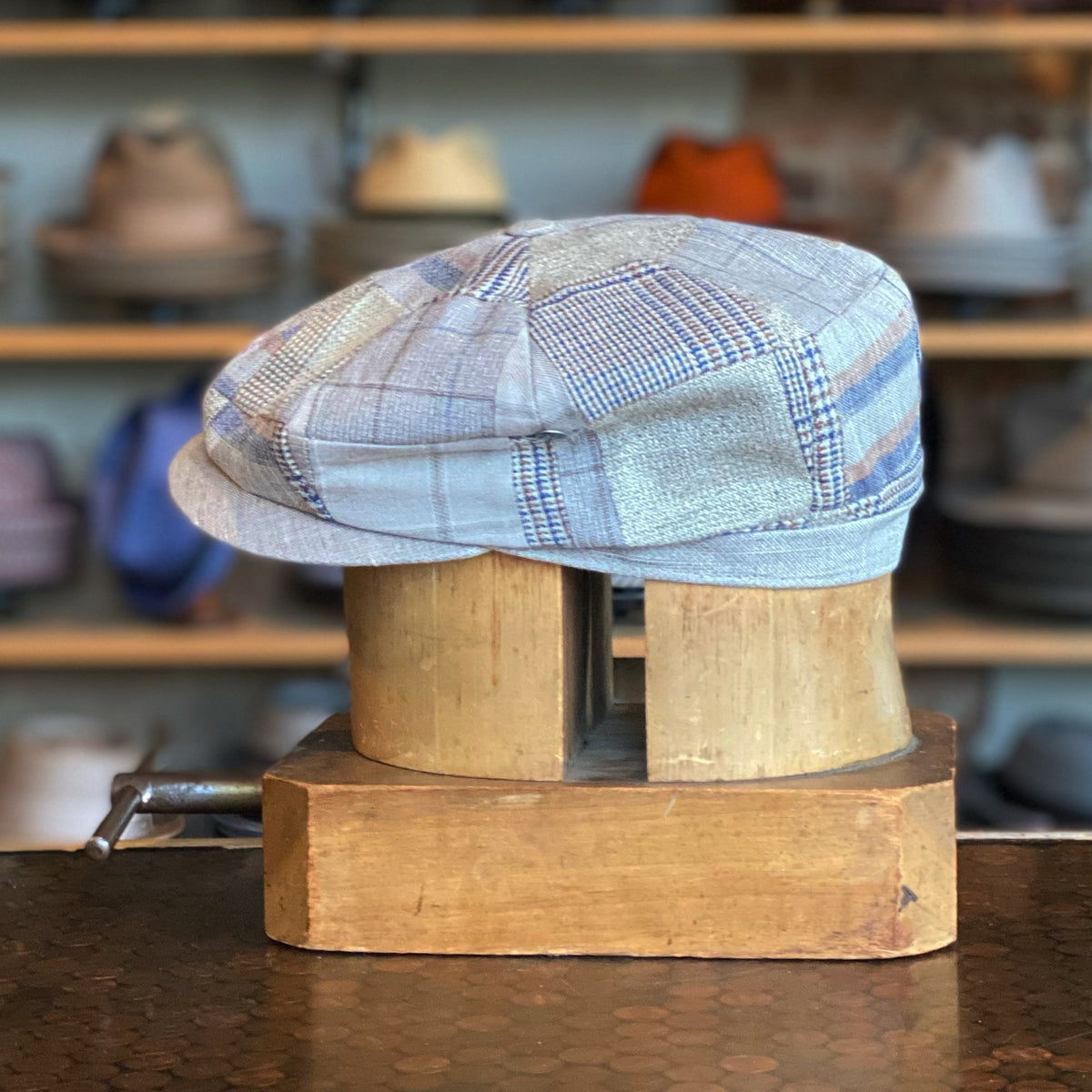 Pattern Patch Hatteras newsboy cap in a soft neutral patchwork fabric displayed on a wooden hat block with shelves of other hats in the background.