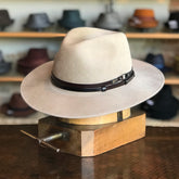 Stedman beige wide-brimmed hat with a dark leather band displayed on a wooden hat block in a shop with various hats blurred in the background