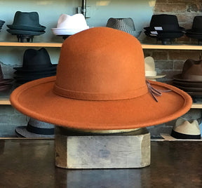 Sundance rust-colored wide-brim felt hat with a subtle decorative bow, displayed on a wooden block in a shop with various other hats in the background.