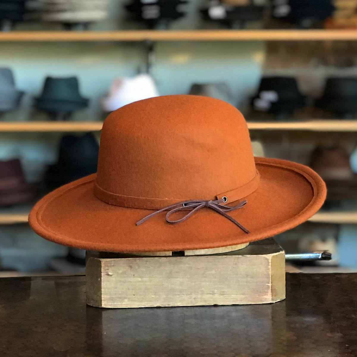 Sundance rust-colored felt hat with a wide brim and thin leather band tied in a bow, displayed on a wooden block with other hats in the background.