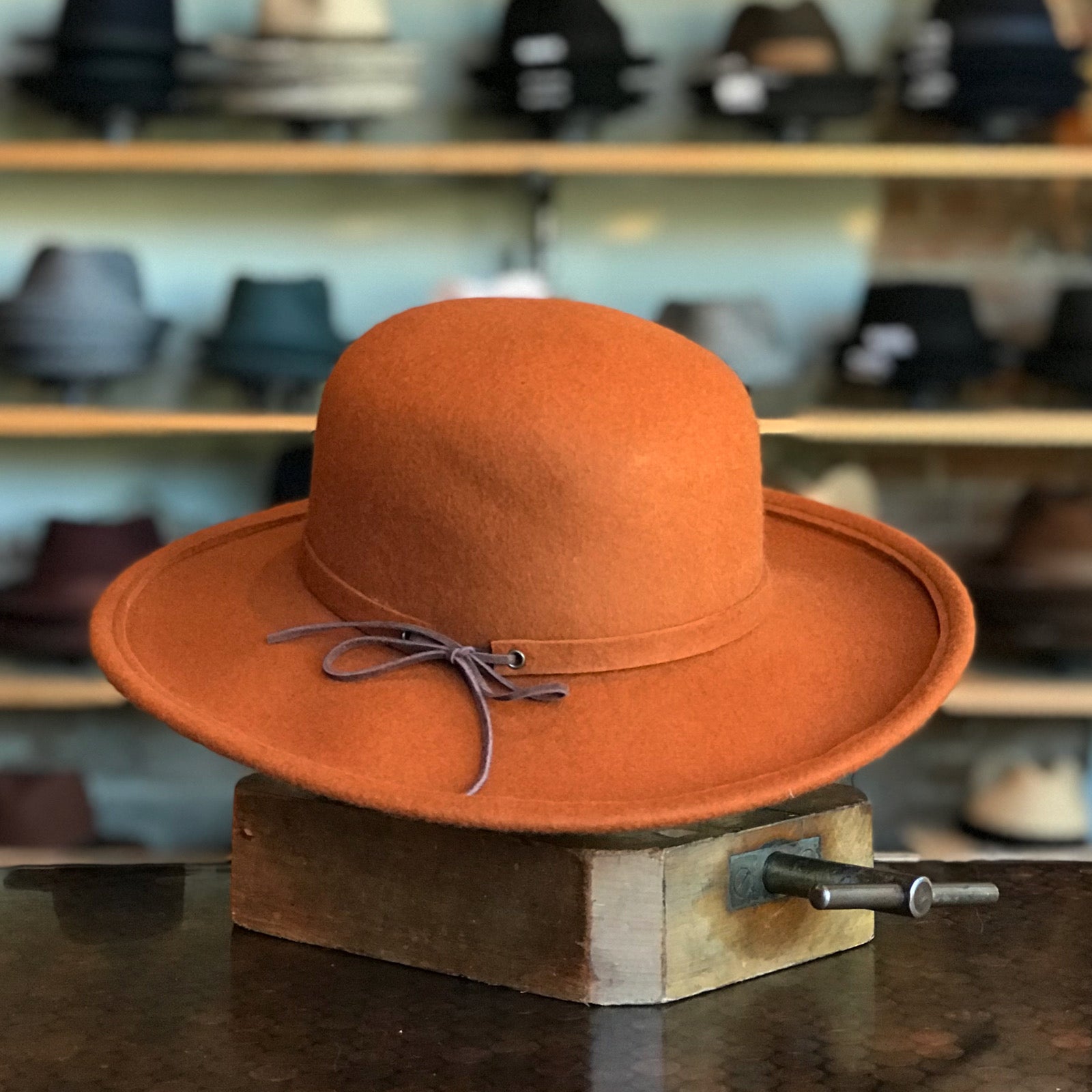 Sundance hat in warm rust color featuring a wide brim and a delicate leather tie detail, displayed in a hat shop with various styles in the background.