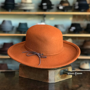 Sundance hat in warm rust color featuring a wide brim and a delicate leather tie detail, displayed in a hat shop with various styles in the background.