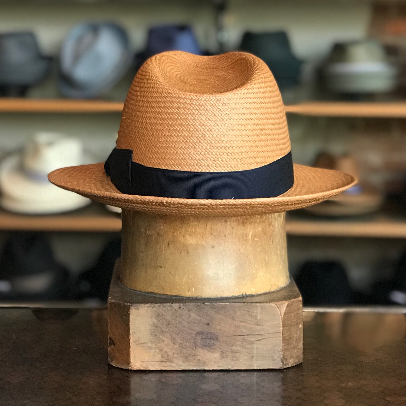 Torino straw hat with a black band displayed on a wooden hat block, set against a background of various hats on shelves in a store.