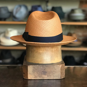 Torino straw hat with a black band displayed on a wooden hat block, set against a background of various hats on shelves in a store.