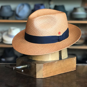 Torino straw fedora hat with a black band and a small red accent, displayed on a wooden hat block with shelves of various hats in the background.