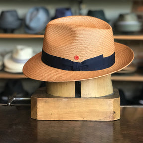 Torino light brown woven hat with a black ribbon band and a small red button detail, displayed on a wooden block with various hats blurred in the background.