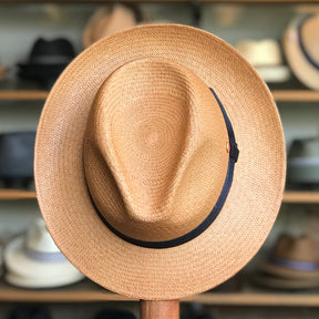 Torino straw hat with a wide brim and black band, displayed in front of a blurred background of various hats on wooden shelves.