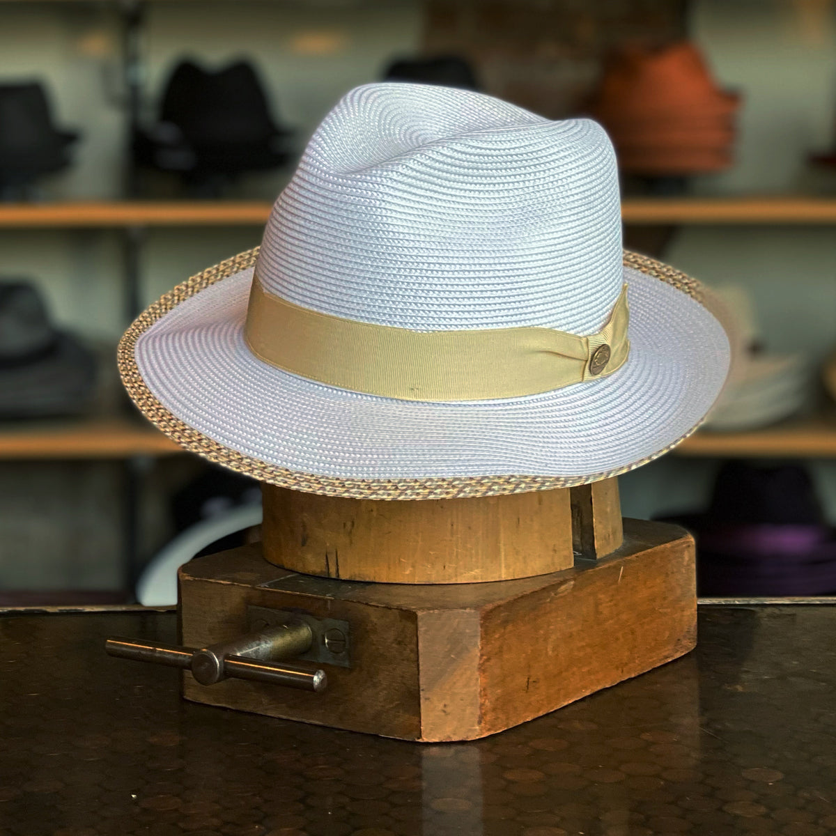 Vidora white woven fedora hat with a beige ribbon band and a braided brim edge, displayed on a wooden hat block in a hat shop setting.