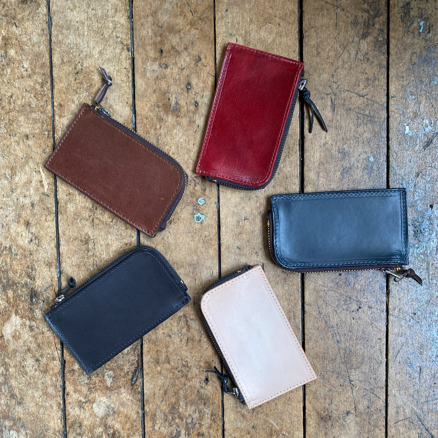 Five leather Zipper Wallets in red, brown, black, navy, and natural tones arranged on a wooden surface.